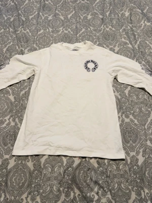 Vit långärmad t-shirt med tryck - Säljer en vit långärmad t-shirt med svart grafiskt tryck på bröstet och längs ena ärmen. T-shirten har en avslappnad passform och är tillverkad i mjuk bomull. Perfekt för dig som gillar unika detaljer och stilrena färger.