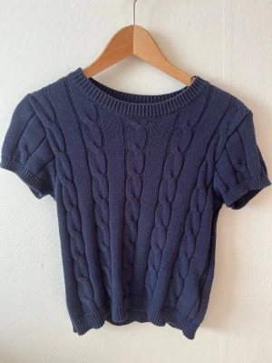 Mörkblå kabelstickad topp Gina Tricot - Kortärmad mörkblå kabelstickad topp från Gina Tricot. Tröjan har rund halsringning, ribbade muddar och ett klassiskt flätmönster över hela plagget.
