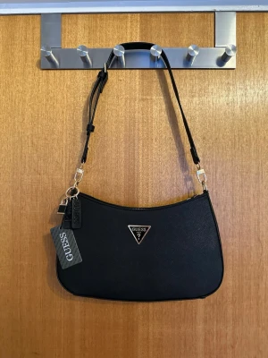 Svart axelremsväska från Guess - Snygg svart handväska från Guess med justerbar axelrem och guldfärgade detaljer. Väskan har Guess-logga i metall framtill och dragkedja upptill. Tillverkad i ett tåligt konstläder med struktur. Perfekt för dig som vill ha en stilren och trendig accessoar.