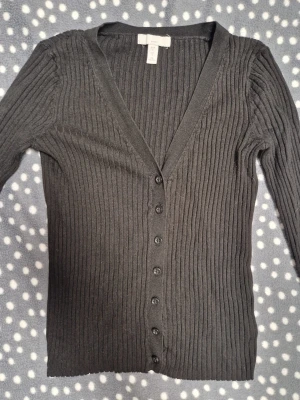 Svart ribbad kofta från H&M - Säljer en svart ribbstickad kofta från H&M med djup V-ringning och knäppning framtill. Koftan har långa ärmar och är tillverkad i ett mjukt, stretchigt material som sitter snyggt på kroppen. Perfekt att slänga över en topp eller t-shirt.