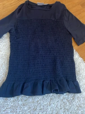 Mörkblå blus med volang från Gina Tricot - Supersöt mörkblå blus från Gina Tricot med smockad midja och volang nertill. Perfekt att styla med jeans eller kjol för en trendig look. Blusen har en enkel rund halsringning och är tillverkad i ett mjukt tyg. I storlek 146/152