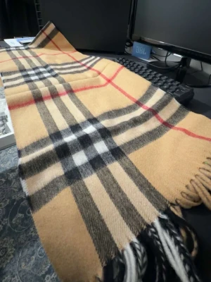  halsduk Burberry - Klassisk beige halsduk från Burberry med det ikoniska rutmönstret i svart, vitt och rött. Halsduken har fransar i ändarna och är tillverkad i mjukt material som känns lyxigt och varmt. Perfekt accessoar för att lyfta din outfit.