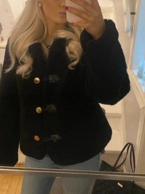 Svart teddyjacka från Zara - Mysig svart teddyjacka från Zara med tre dekorativa guldknappar och tre svarta spännen framtill. Jackan har en klassisk krage och är tillverkad i fluffigt teddy-material som ger en trendig look. Perfekt för att hålla värmen under kyliga dagar.