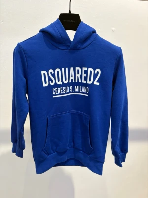 Blå hoodie från Dsquared2 - Blå hoodie med vit Dsquared2-logga och texten 'Ceresio 9, Milano' på bröstet. Klassisk känguruficka framtill och huva. Perfekt för en avslappnad och trendig stil.