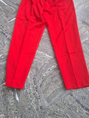 Röda kostymbyxor med knytskärp - Säljer ett par snygga röda kostymbyxor med hög midja och tillhörande knytskärp i midjan. Byxorna har raka ben och sidofickor för en clean look. Perfekta för dig som vill sticka ut med färg och stil.