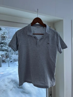 Ljusgrå pikétröja från Gant - Snygg ljusgrå pikétröja från Gant med klassisk krage och två knappar vid halsen. Tröjan har korta ärmar och en liten broderad logga på bröstet. Perfekt för en avslappnad men stilren look.