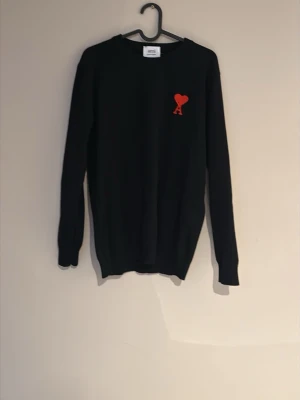 Svart stickad tröja från Ami - Svart stickad tröja från Ami Alexandre Mattiussi med röd broderad logga i form av ett hjärta och bokstaven A på bröstet. Tröjan har rund halsringning och långa ärmar. Perfekt för dig som gillar stilrena plagg med en twist.