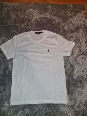 Vit t-shirt från Ralph Lauren - Klassisk vit t-shirt från Ralph Lauren med den ikoniska broderade polospelaren på bröstet. T-shirten har rund halsringning och korta ärmar. Perfekt basplagg som funkar till allt och är tillverkad i mjuk bomull för skön känsla.