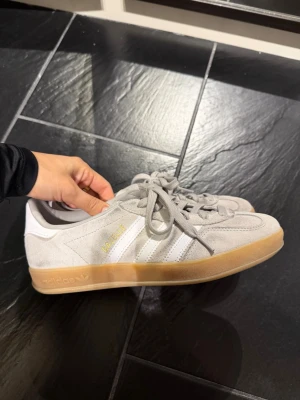 Adidas Gazelle ljusgrå sneakers - Säljer ett par Adidas Gazelle sneakers i ljusgrå mocka med vita detaljer och klassiska tre ränder på sidan. Skorna har gummisula i beige och guldfärgad Gazelle-text på sidan. Perfekta för en clean och stilren look.