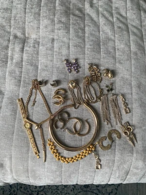 Mix av guldfärgade smycken - Säljer ett blandat paket med guldfärgade smycken, inklusive armband, halsband, örhängen, ringar och en klocka. Några smycken har detaljer i lila och pärlor. Perfekt för dig som gillar att variera din stil med olika accessoarer.