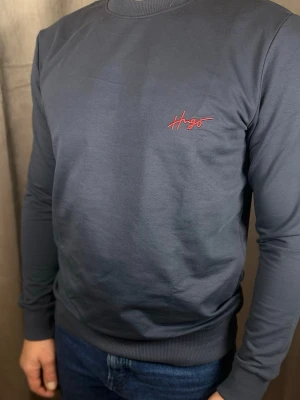 Mörkgrå stickad tröja från Hugo - Snygg mörkgrå stickad tröja från Hugo med röd broderad logga på bröstet. Tröjan har rund halsringning, långa ärmar och ribbade muddar vid ärmslut och nederkant. Perfekt för en stilren och avslappnad look.