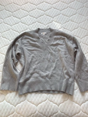 Grå stickad v-ringad tröja - En stilren grå stickad tröja med v-ringning och breda ribbade muddar vid ärmslut och nederkant. Perfekt för lager-på-lager och enkel att matcha med jeans eller kjol. Tröjan har en klassisk passform och är långärmad.