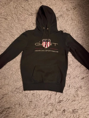 Svart hoodie från gant  - Svart hoodie från gant med huva och dragsnören. Storlek S, På bröstet finns ett stort tryck med GANT-logga och texten 'American Sportswear' samt ett sköldmärke i rött, vitt och guld. Klassisk känguruficka framtill och ribbade muddar. Perfekt för en sportig och avslappnad stil. Ny pris 1200kr!