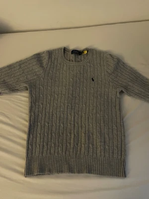 Grå kabelstickad tröja från Polo Ralph Lauren - Kabelstickad grå tröja från Polo Ralph Lauren med rund halsringning och broderad logga på bröstet. Tröjan har långa ärmar och ribbstickade muddar vid ärmslut och nederkant. Perfekt för lager-på-lager och klassisk stil.