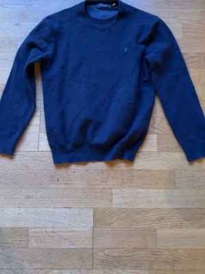 Mörkblå tröja från Polo Ralph Lauren - Klassisk mörkblå tröja från Polo Ralph Lauren med rund halsringning och diskret broderad logga på bröstet. Tröjan har långa ärmar och ribbade muddar vid ärmslut och nederkant. Perfekt för en stilren och enkel look.