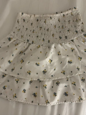 Vit blommig volangkjol Gina Tricot - Supersöt kort vit kjol från Gina Tricot med små blommor i blått och gult. Kjolen har bred smock i midjan och dubbla volanger för en extra flowy look. Perfekt för sommarens alla tillfällen!