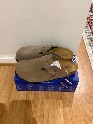 Birkenstock Boston  - Snygga beige tofflor från Birkenstock i mjuk mocka med klassisk design och justerbart spänne i metall. Fotbädden är formad för extra komfort och sulan är i kork och gummi. Perfekta för dig som vill ha både stil och bekvämlighet.