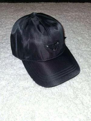 Svart keps från Prada i nylon - Snygg svart keps från Prada i glansigt nylonmaterial med klassisk böjd skärm. Framtill sitter Pradas ikoniska trekantiga metallogo. Perfekt accessoar för en trendig och stilren look.