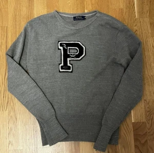 Stickad Ralph Lauren Tröja - Grå stickad tröja från Polo Ralph Lauren med svartvit P-applikation. Klassisk college-stil med rund hals och ribbade muddar. Midjan har krympt något – perfekt för slim fit och passar även Small.