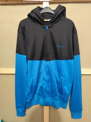 Svart och blå hoodie från Carhartt - Snygg hoodie från Carhartt i svart och blått med dragkedja framtill och två fickor med blå dragkedjor. Broderad Carhartt-logga i blått på bröstet. Tillverkad i ett mjukt och bekvämt material, perfekt för en avslappnad stil. Huva med dragsko för extra komfort.