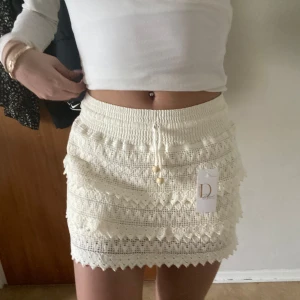 Vit virkad minikjol med knytband - Beige kjol Supersöt vit virkad minikjol från Demoiselle.se  med knytband och små pärldetaljer framtill. Kjolen har ett fint mönster och elastisk midja för skön passform. Perfekt till varma dagar och enkel att styla med olika toppar.