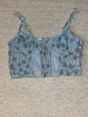 Blå spets corset med blommönster - Säljer en blå corset med broderade blommor i ljusblått och mörkblått. Corseten har justerbara axelband och är tillverkad i genomskinlig mesh och spets.