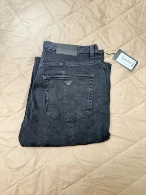Svarta jeans från Emporio Armani - Snygga svarta jeans från Emporio Armani med klassisk femficksmodell och logga broderad på bakfickan. Jeansen har raka ben och är tillverkade i mjuk denim med en modern tvätt. Perfekta för en trendig och avslappnad stil.