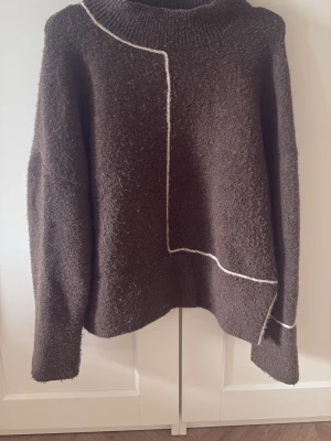 Brun stickad tröja med vit detalj - Mysig brun stickad tröja med unika vita linjer som skapar ett grafiskt mönster. Tröjan har en hög, ribbstickad krage och långa ärmar. Perfekt för dig som gillar en stilren men ändå trendig look.