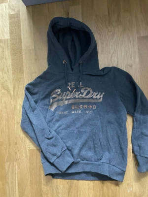 Grå Superdry hoodie med tryck - Mysig blå hoodie från Superdry med metallicfärgat logotryck på bröstet och japanska tecken. Klassisk känguruficka framtill och justerbar huva med snören. Perfekt för chill dagar och streetwear-stil. 