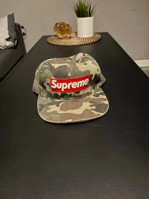 Camo Supreme keps med röd logga - Säljer en camouflagemönstrad keps från Supreme med den klassiska röda box loggan framtill. Kepsen har platt skärm och är tillverkad i slitstarkt tyg, perfekt för dig som vill sticka ut med streetwear-stil.