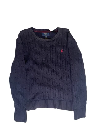 Mörkblå kabelstickad tröja Polo Ralph Lauren - Kabelstickad mörkblå tröja från Polo Ralph Lauren med rund halsringning och röd broderad logga på bröstet. Tröjan har ribbade muddar vid ärmslut och nederkant. Perfekt för en klassisk och stilren look.obs lite solblekt 