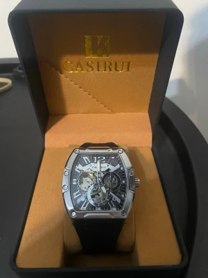 CASIRUI silverfärgad klocka - Snygg klocka från CASIRUI med fyrkantig silverfärgad boett och svart urtavla med synliga mekaniska detaljer. Klockan har svarta band och levereras i originalask. Perfekt accessoar för dig som gillar stilrena och moderna detaljer.