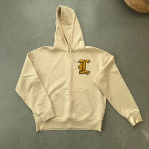 Lacoste Beige hoodie - Snygg beige hoodie från Lacoste med stor gul broderad L-logga på bröstet.  Klassisk huva med dragsko och magficka framtill. Perfekt för en avslappnad och trendig stil. Köpt några år sedan och inte använd på något år. OBS se fläck på sista bilden. 