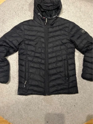 Svart dunjacka med huva - Snygg svart dunjacka med huva och dragkedja framtill. Jackan har två sidofickor med dragkedja och quiltad design. Perfekt för kalla vinterdagar och riktigt lätt att bära.