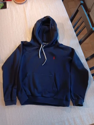 Mörkblå hoodie från Polo Ralph Lauren - Snygg mörkblå hoodie från Polo Ralph Lauren med klassisk röd logga broderad på bröstet. Hoodien har en stor huva med vita dragsnören och en rymlig magficka framtill. Tillverkad i mjukt bomullsmaterial och har ribbade muddar vid ärmar och nederkant.