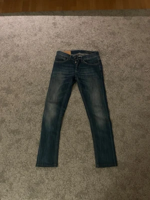 Mörkblå dondups - Snygga mörkblå dondups med klassisk femficksdesign och diskreta slitningar på låren. Jeansen har normal midja och raka ben, perfekta för en avslappnad men stilren look. Passar till det mesta och är ett måste i garderoben. Dem är 39 cm i midjan och 101 cm på längden! Hör av er vid intresse!