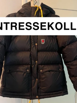 Svart dunjacka från Fjällräven - INTRESSEKOLL! Svart dunjacka från Fjällräven med huva, gula snören och tryckknappar. Jackan har två stora fickor framtill och klassisk Fjällräven-logga på bröstet. Puffig modell som håller dig varm under kalla dagar. OBS! Finns ett lagat hål på jackan(se bild 4). Den har används i två vintrar och kan tänka mig att sälja den till en seriös köpare .Skriv gärna ifall ni är intresserade av att köpa för ungefär det priset som jag har satt.  Fjällräven påsen följer med vid köpet. 
