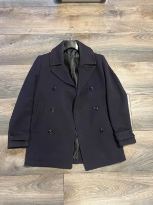 Mörkblå dubbelknäppt rock - Stilren mörkblå rock med dubbelknäppning och bred krage. Rocken har två sidofickor, dekorativa knappar vid ärmsluten och helsvart innerfoder. Klassisk och tidlös modell som passar perfekt till höst och vinter.