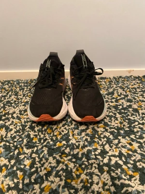 Svarta asics gel nimbus originalpris 2200kr - Snygga svarta sneakers från Asics med klassiska ränder i orange och vitt på sidorna. Skorna har tjock vit sula med orange inslag framtill och är tillverkade i mesh och syntetmaterial. Perfekta för dig som gillar sportig stil och vill ha bekväma skor till vardags.