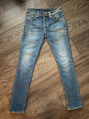  Nudie grim tim - Snygga blå Nudie grim Tim Jeans med klassisk femficksdesign och slitningar framtill. Jeansen har orangea sömmar och broderade bakfickor. Tillverkade i bomull med en lätt stretch för extra komfort. Där är en liten reva/hål vid låret som man ser på sista bilden men syns typ bara på nära håll. Annars bra skick