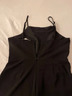 Svart jumpsuit med smala axelband - Snygg svart jumpsuit med smala axelband och raka, vida ben. Enkel och stilren design som passar perfekt till många tillfällen. Jumpsuiten har en figurnära överdel och är tillverkad i ett mjukt tyg som ger en bekväm känsla.