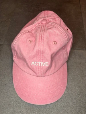 Rosa keps från GAAM med brodyr - Säljer en rosa keps från GAAM med vit brodyr framtill där det står 'ACTIVE' och bak med GAAM-loggan. Klassisk baseballmodell med böjd skärm och justerbar rem bak. Tillverkad i bomullstyg och har ventilationshål för extra komfort.