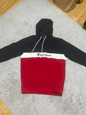Champion jacka svart/röd med huva - Snygg hoodie från Champion i svart och rött med vit panel och broderad logga över bröstet. Klassisk huva med vita snören och känguruficka framtill. Perfekt för en avslappnad streetwear-look.