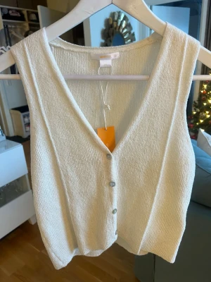 Vit stickad väst med knappar - Snygg vit stickad väst med v-ringning och knappar framtill. Perfekt att styla över en t-shirt eller skjorta för en trendig lager-på-lager-look. Mjuk och bekväm med en enkel, clean design.