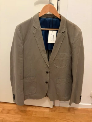 Beige kavaj från Paul Smith - Stilren beige kavaj från Paul Smith med två knappar framtill, tre utanpåfickor och klassisk krage. Kavajen har blått innerfoder och är tillverkad i ett lätt bomullstyg, perfekt för en snygg och avslappnad look.