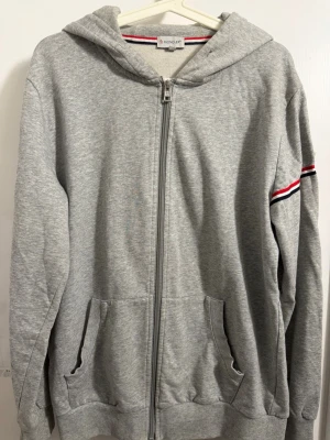 Grå zip hoodie från Moncler. Bara att skriva om du har några frågor. - Snygg grå hoodie från Moncler med dragkedja framtill och två fickor. Klassisk huva och ribbade muddar. Ärmen har en röd, vit och blå rand samt Moncler-logga. Tillverkad i mjukt bomullsmaterial som känns skönt mot huden. Men passar m och L oxå.