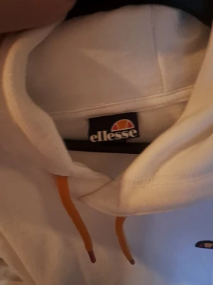 Vit hoodie från Ellesse med orange snören - Säljer en vit hoodie från Ellesse med orange dragsnören och liten broderad logga på bröstet. Hoodien har en klassisk känguruficka framtill och ribbade muddar. Perfekt för dig som gillar sportig och clean stil.