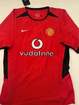 Sprillans Ny Manchester United Matchtröja Ferdinand #5 - Klassisk röd Manchester United matchtröja från Nike med svart detalj, klubbmärke på bröstet och Vodafone som sponsor. På ryggen står det 'Ferdinand' och nummer 5. Perfekt för fotbollsfans och samlare.
