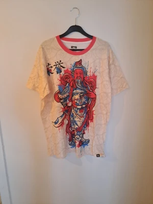 Beige t-shirt med asiatisk print - Cool beige t-shirt med stort färgglatt asiatiskt motiv i rött och blått framtill. T-shirten har korta ärmar, rund hals med röd kant och är tillverkad i mjuk bomull. Perfekt för dig som vill sticka ut med en unik stil. Liten I storlek som en xl