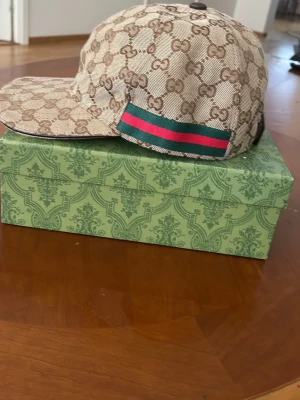 Gucci keps med monogram och ränder - Snygg keps från Gucci i beige canvas med klassiskt GG-monogram över hela kepsen. På sidan finns de ikoniska gröna och röda ränderna. Justerbar rem baktill i mörkbrunt läder. Perfekt accessoar för en trendig streetstyle.